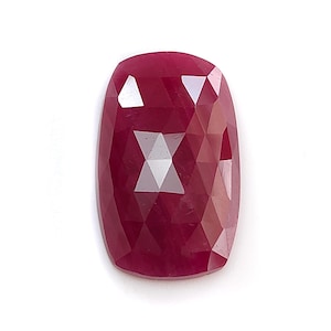 May include: A deep red, cushion-cut gemstone with a faceted surface. The stone features a rectangular shape with rounded corners and a polished finish that reflects light across its multiple triangular facets, showcasing a vibrant, saturated colour.