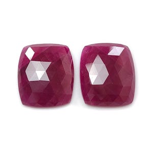 May include: Two faceted, cushion-cut rubies. The gems are a deep, rich red color with multiple facets that reflect light. The rubies are set against a white background, highlighting their color and clarity. The Saksham Gems logo is in the top center.