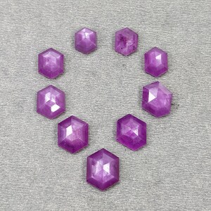 May include: Nine hexagonal gemstones in shades of purple are arranged in a circle on a grey surface. The faceted stones have a polished, reflective surface. The gemstones are suitable for jewellery making.