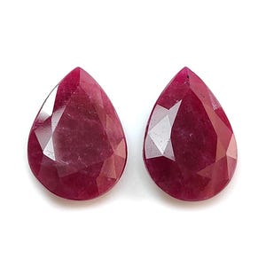 May include: Two pear-shaped, faceted rubies with a deep, saturated magenta-red colour. Each gemstone features a pointed top and a rounded base, displaying a polished surface with geometric facets that reflect light across the translucent stone.