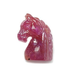 May include: A carved gemstone sculpture of a horse's head. The sculpture is a vibrant pink colour with intricate details, showcasing the mane and facial features. The horse head is set against a white background.