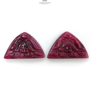 Könnte beinhalten: Zwei dreieckige, tiefrote geschnitzte Edelsteine mit einem detaillierten Blumenmotiv auf ihren Oberflächen. Die Steine haben eine polierte Oberfläche und sind nebeneinander angeordnet. Das Logo SAKSHAM GEMS THE EFFICACY ist oben sichtbar.