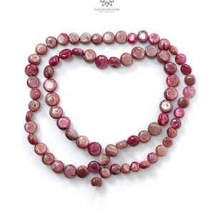 Può includere: Un filo di perline di pietre preziose rotonde e piatte, di colore rosso rosato, disposte a forma di cuore. Le perline presentano una superficie lucida e leggermente irregolare con varie tonalità di magenta intenso e rosa chiaro. Il logo Saksham Gems è in alto.