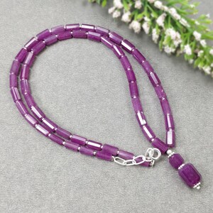 Könnte beinhalten: Eine Halskette aus lila Edelsteinperlen mit einem silbernen Verschluss. Die Kette besteht aus kleinen, rechteckigen Perlen und hat einen passenden Anhänger. Die Halskette wird auf einer grauen Oberfläche präsentiert.