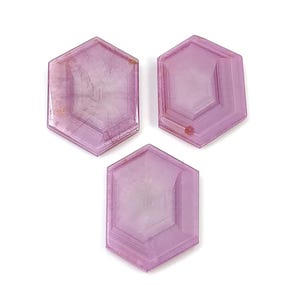 Könnte beinhalten: Drei sechseckige, facettierte Edelsteine in verschiedenen Rosa- und Lilatönen werden vor einem weißen Hintergrund gezeigt. Die Edelsteine haben eine durchscheinende Qualität mit sichtbaren inneren Mustern und einer polierten Oberfläche. Die Anordnung ist optisch ansprechend.