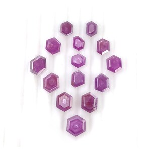 May include: A collection of thirteen loose, faceted gemstones in a vibrant magenta-pink hue. Each stone features a distinct hexagonal shape with polished surfaces that reflect light, arranged in a symmetrical pattern against a plain white background.