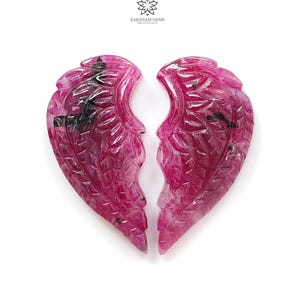 Puede incluir: Dos alas talladas en piedras preciosas de rubí, cada una con intrincados detalles de plumas. Las alas son de un rosa vibrante con inclusiones oscuras, sobre un fondo blanco. El logotipo "SAKSHAM GEMS THE ETHICALS" está en la parte superior.