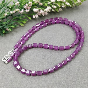 Puede incluir: Un collar con piedras preciosas facetadas de color púrpura intenso, ensartadas con pequeñas cuentas plateadas. El collar se exhibe sobre una superficie gris, con una ramita de vegetación y pequeñas flores blancas en el fondo.