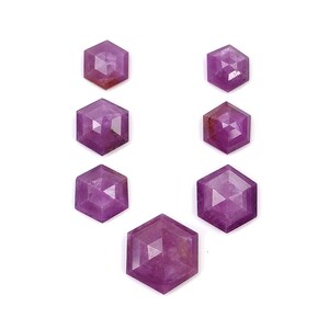 Puede incluir: Una colección de siete piedras preciosas sueltas y facetadas en forma hexagonal. Cada piedra presenta un tono magenta-púrpura vibrante con una superficie geométrica pulida que refleja la luz. Las piedras están dispuestas en forma de U.