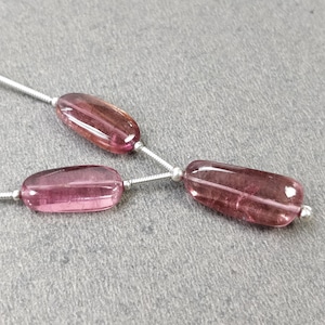 May include: Three pink and purple tourmaline gemstone beads on a silver string. The beads are oblong and translucent, with variations in color. The beads are strung together, creating a simple, elegant design. The background is a neutral gray.