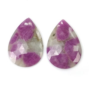 Może przedstawiać: Dwa fasetowane kamienie szlachetne w kształcie łezki. Kamienie mają mieszankę głębokich różowych i białych kolorów. Różowe sekcje mają krystaliczny wygląd, podczas gdy białe obszary są bardziej nieprzezroczyste. Kamienie są na białym tle.