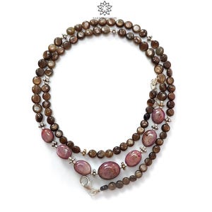 May include: A layered necklace featuring brown and pink gemstones. The necklace has three strands of small, round, brown beads, interspersed with larger, oval, pink gemstones. The necklace is secured with a silver clasp.