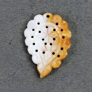 May include: A two-toned pendant with a floral design. The pendant is shaped like a leaf, with a white, scalloped edge on one side and a golden-orange, scalloped edge on the other. Small holes are drilled throughout the pendant.