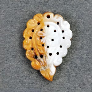 May include: A carved pendant with a floral design, featuring a combination of orange and white colors. The pendant has multiple small holes and a scalloped edge. The overall shape is reminiscent of a stylized leaf or flower.