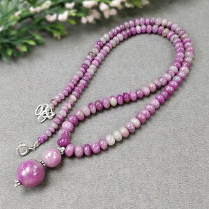 May include: A necklace featuring a strand of small, round, pink and purple beads. The necklace has a larger, round pendant bead and a silver clasp. The beads vary in color from light pink to deep purple.