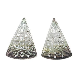 May include: Two pearlescent, triangular pendants. Each pendant has a carved floral design with swirling patterns and small, circular cutouts. The edges are detailed with a textured border. The pendants are displayed against a white background.
