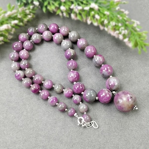 May include: A necklace featuring round, polished beads in shades of purple and gray. The beads are strung together with a silver clasp. The necklace has a large, single bead at the end.
