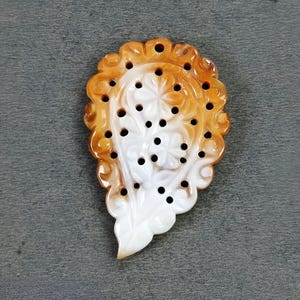 May include: A carved pendant with a floral design. The pendant features a combination of white and orange-brown colors. The edges are scalloped, and there are several small, evenly spaced holes throughout the design.