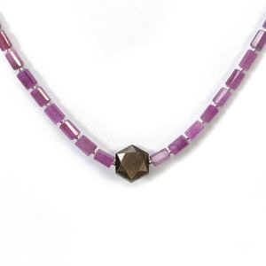 May include: A necklace featuring a central, faceted, dark brown gemstone. The necklace is strung with small, rectangular, pinkish-purple beads, connected by silver-colored links. The necklace is displayed against a white background.