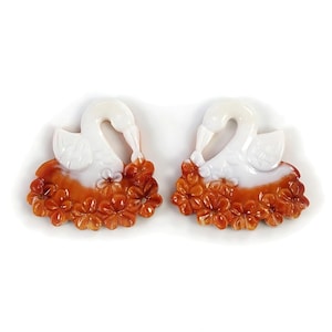 May include: Two swan figurines with white bodies and orange floral bases. The swans have curved necks and are surrounded by detailed orange flowers. The figurines are set against a white background, highlighting their intricate design.