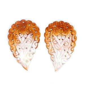 May include: Two ornate, leaf-shaped carvings. The carvings feature a floral design with intricate details and a scalloped edge. The color gradient transitions from a light cream to a warm orange-brown. The carvings are likely decorative elements.