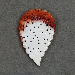 May include: A decorative, leaf-shaped pendant with a white base and a reddish-brown, scalloped top. The white section features a floral design and small, evenly spaced black dots. The pendant is likely made of a polished stone or resin.