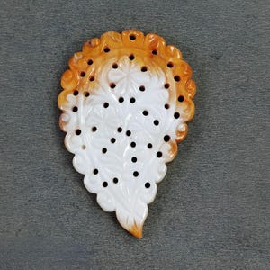 May include: A carved pendant with a leaf-like shape. The pendant features a white base with intricate floral patterns and small black holes. The edges are a warm orange color, creating a striking contrast. The pendant is likely made of stone or a similar material.