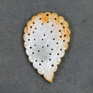 May include: A carved, pear-shaped pendant with a floral design. The pendant is white with a scalloped, amber-colored edge and small, evenly spaced holes. The floral design includes flowers and leaves.