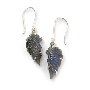 May include: A pair of earrings with wing-shaped pendants. The pendants are dark grey with blue and silver details, suspended from silver-coloured ear wires. The earrings are presented against a white background.