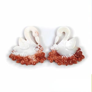 May include: Two white swan figurines with a reddish-brown floral base. The swans have curved necks and are facing each other. The flowers are detailed and add a decorative element to the figurines.