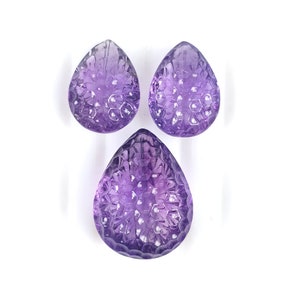 May include: Three carved amethyst gemstones in a teardrop shape. The purple stones have a detailed floral pattern carved into their surface. The gemstones are displayed on a white background.