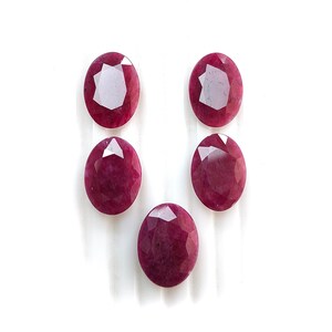 May include: Five oval-cut, faceted ruby gemstones. The rubies are a deep, rich red color and are arranged against a white background. The gemstones have a polished, reflective surface.