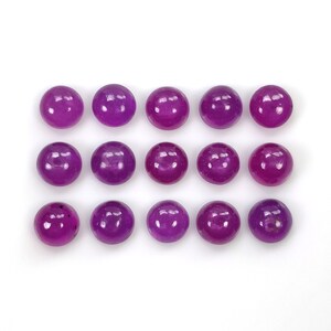 Puede incluir: Quince cabujones redondos, lisos y pulidos de un vibrante color púrpura magenta. Las piedras están dispuestas en una cuadrícula de tres filas y cinco columnas, cada una con una superficie brillante y reflectante que capta la luz con reflejos blancos.