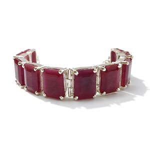 Puede incluir: Una pulsera que presenta una serie de piedras preciosas rectangulares, de color rojo intenso y opacas, engastadas en garras de metal plateado. Las piedras están dispuestas en cadena, cada una sujeta por cuatro garras metálicas en las esquinas.
