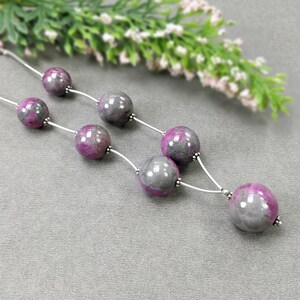 May include: A necklace featuring large, round beads in shades of gray and magenta. The beads are strung on a thin, silver-colored chain. The necklace is displayed on a gray surface, with a blurred floral element in the background.