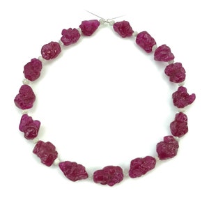 Puede incluir: Un collar que presenta una serie de piedras de rubí en bruto de color rosa intenso dispuestas en un patrón circular. Cada rubí está separado por una pequeña cuenta blanca translúcida y texturizada. Las piedras tienen una forma irregular.