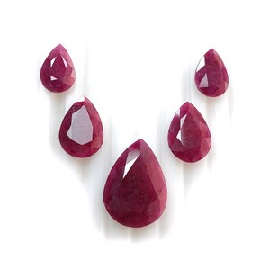 Peut inclure: Cinq rubis facettés en forme de poire disposés en demi-cercle. Les pierres précieuses présentent une couleur rouge magenta saturée avec des inclusions internes visibles et des facettes géométriques nettes qui reflètent la lumière.