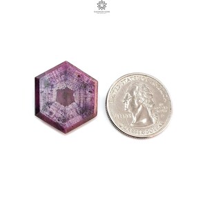 Pode incluir: Uma pedra preciosa hexagonal e facetada, de cor rosa profundo, com padr&otilde;es internos, ao lado de uma moeda de 25 centavos americana para compara&ccedil;&atilde;o de tamanho. A pedra tem uma &aacute;rea central mais clara e bordas mais escuras.