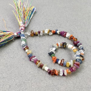 May include: A strand of colorful gemstone beads with a multi-colored tassel. The beads are a mix of red, orange, yellow, green, blue, purple, and white. The beads are strung on a clear thread.