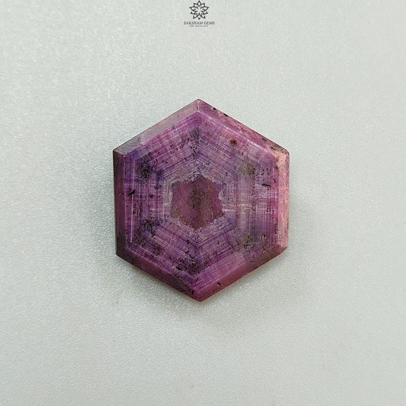Pode incluir: Uma pedra preciosa hexagonal polida, com um tom rosa profundo e um padr&atilde;o em forma de estrela no centro. A gema tem uma superf&iacute;cie facetada e uma apar&ecirc;ncia natural e texturizada. Est&aacute; sobre um fundo branco.