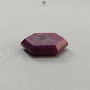 May include: A faceted, hexagonal gemstone with a deep magenta hue. The gem has a polished surface and visible internal patterns. The stone is set against a neutral background, highlighting its color and shape. The gem is likely a ruby.