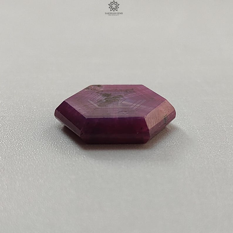 May include: A faceted, hexagonal gemstone with a deep magenta hue. The gem has a polished surface and visible internal patterns. The stone is set against a neutral background, highlighting its color and shape. The gem is likely a ruby.