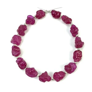 Puede incluir: Un collar de piedras preciosas de rubí magenta en bruto. Cada piedra tiene una forma irregular y está ensartada con pequeñas cuentas de color plateado. Las piedras preciosas se muestran sobre un fondo blanco brillante.