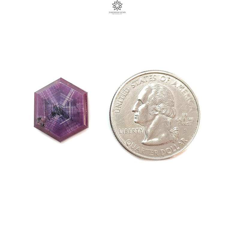 May include: A faceted, hexagonal gemstone with a deep purple hue, displayed next to a U.S. quarter for size comparison. The gem has internal patterns and inclusions, set against a white background. The gem is approximately 0.75 inches in diameter.
