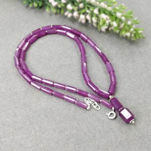May include: A necklace made of purple gemstones with a silver clasp. The necklace is composed of small, rectangular beads, with a larger rectangular pendant. The beads are a deep purple colour.