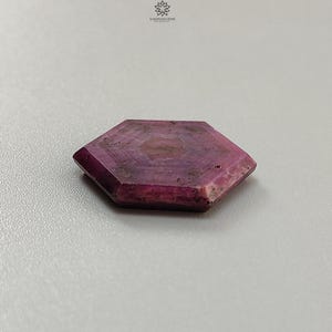 Pode incluir: Uma pedra preciosa de rubi hexagonal e facetada. A gema &eacute; de cor vermelho-p&uacute;rpura profunda com padr&otilde;es internos mais claros. As bordas s&atilde;o bem definidas e a pedra tem uma textura ligeiramente &aacute;spera. A pedra preciosa &eacute; exibida em uma superf&iacute;cie cinza claro.