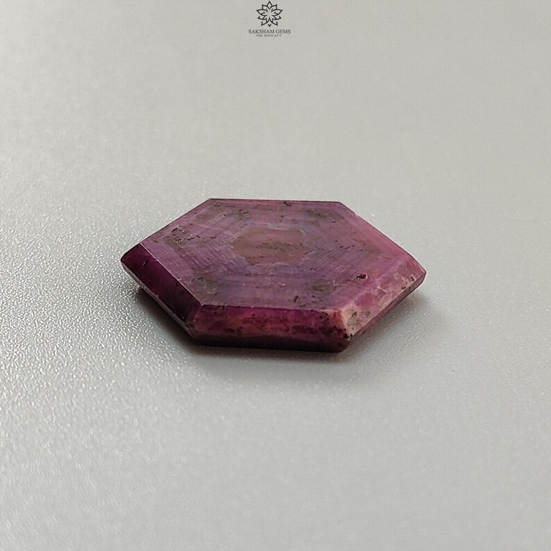 Pode incluir: Uma pedra preciosa de rubi hexagonal e facetada. A gema &eacute; de cor vermelho-p&uacute;rpura profunda com padr&otilde;es internos mais claros. As bordas s&atilde;o bem definidas e a pedra tem uma textura ligeiramente &aacute;spera. A pedra preciosa &eacute; exibida em uma superf&iacute;cie cinza claro.