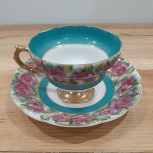 May include: A vintage teacup and saucer set with a floral design. The cup and saucer are white with a turquoise blue rim and a gold trim. The floral design features pink roses and green leaves.