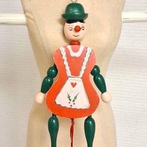 May include: A vintage wooden pull-string doll with a green hat, red nose, and orange dress. The doll has articulated limbs and a white apron with a heart and holly design. The doll is suspended by a red string.