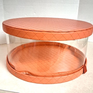 May include: A clear, round display case with a peach-colored, quilted top and base. The sides are transparent, allowing a full view of the contents. The case has a small, peach-colored tab on the side. The case is designed for displaying items.